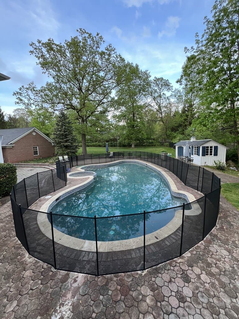 Kidney pool with removable mesh safety fence, clear water, and patterned pavers, typical finish after leak checks, coping resets, and seasonal startup work.
