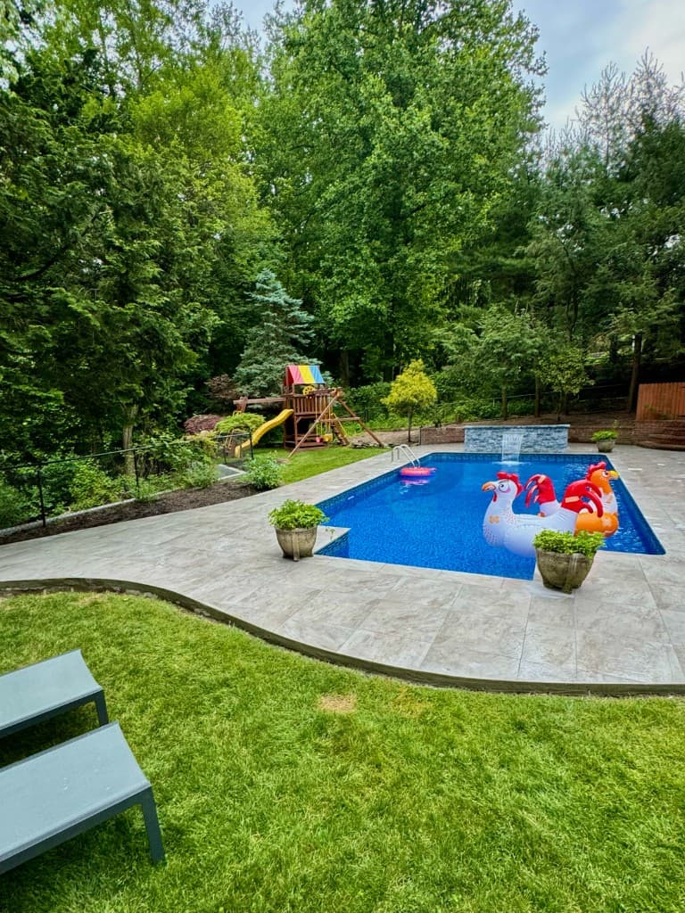 Finished backyard pool with clear blue water, stone patio, sheer-descent water feature, floats, and wooded backdrop after Shiny Pools service